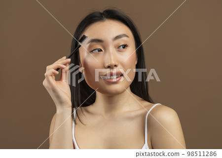 Beautiful young japanese lady brushing brows and applying eyebrow mascara, posing over brown studio background Beautiful young japanese lady brushing brows and applying eyebrow mascara, posing over brown studio background 98511286