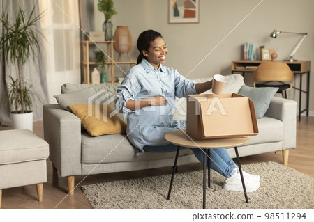 Happy young pregnant black lady unpacking box and holding cup, touching belly and smiling, sitting on sofa at home 98511294