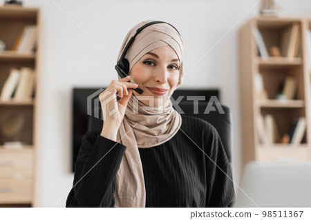 Smiling positive confident woman in hijab and headset sitting at office desk. 98511367