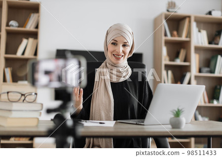 Smiling arab woman in beige hijab sitting at desk with books, notes and laptop recording video Smiling arab woman in beige hijab sitting at desk with books, notes and laptop recording video 98511433