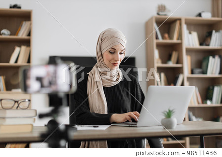 Confident muslim female in headscarf sitting at desk working on laptop and recording video Confident muslim female in headscarf sitting at desk working on laptop and recording video 98511436