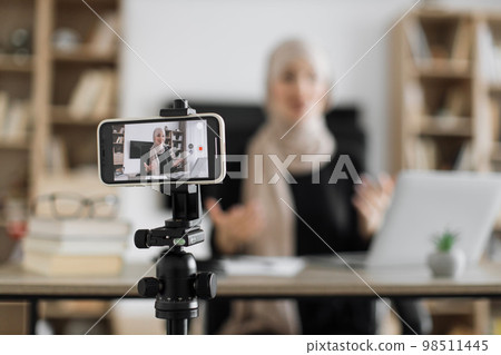Blur background of pretty muslim woman with headscarf sitting at desk and filming video blog. 98511445