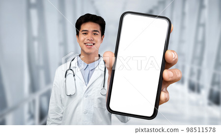 Smiling Asian Male Doctor Wearing Uniform Showing Big Smartphone With Blank Screen Smiling Asian Male Doctor Wearing Uniform Showing Big Smartphone With Blank Screen 98511578