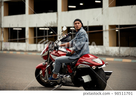 Lifestyle of a motorcyclist, a handsome biker with long hair on a red classic bike in an urban landscape. Lifestyle of a motorcyclist, a handsome biker with long hair on a red classic bike in an urban landscape. 98511701