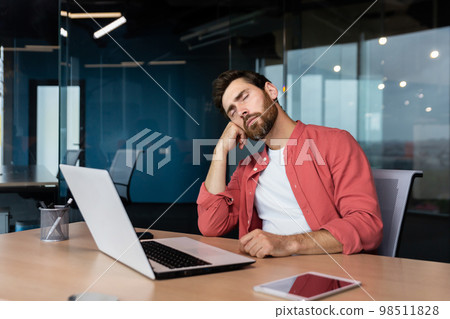 Tired businessman in casual red shirt sleeps at workplace, man on desk fell asleep during working hours inside office with laptop. 98511828