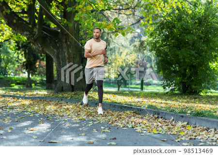 Hispanic man jogging in the park on a sunny day, runner listening to music in wired headphones, audio books and podcasts, sportsman happy with an active weekend. 98513469