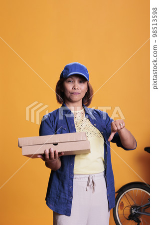 Serious pizzeria delivery worker doing disapproval sign while holding carton cardboard, delivering take away food to customers. Courier bringing pizza orders with bike. Take out food service 98513598