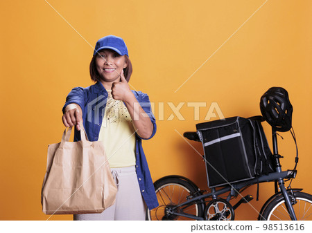 Smiling deliverywoman doing thumbs up gesture while delivering take away food to customers, using paper bags. Restaurant worker bringing orders with bike. Takeout food service and concept 98513616