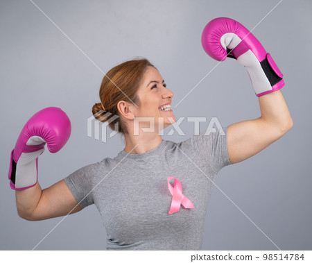 A woman with a pink ribbon on her chest raises her hands in pink boxing gloves on a gray background. Victory over breast cancer. A woman with a pink ribbon on her chest raises her hands in pink boxing gloves on a gray background. Victory over breast cancer. 98514784
