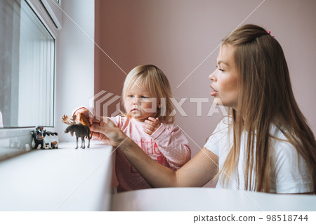 Little girl toddler with her mother playing with animal toys on window sill in children's room at home Little girl toddler with her mother playing with animal toys on window sill in children's room at home 98518744