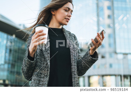 Young smiling woman in coat with coffee cup using mobile phone in evening city street 98519184
