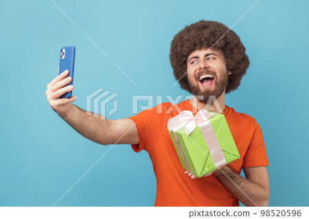 Portrait of happy joyful man with Afro hairstyle wearing orange T-shirt having video call with present box, boasting his gift to followers. Indoor studio shot isolated on blue background. 98520596