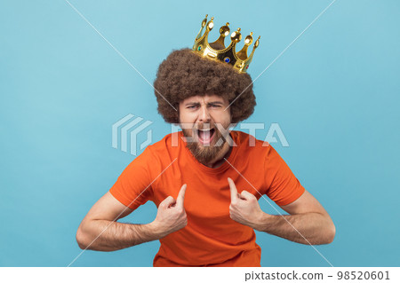 Portrait of crazy man with Afro hairstyle in golden crown and pointing himself with excite expression, looking with arrogance, declaring his authority. Indoor studio shot isolated on blue background. 98520601