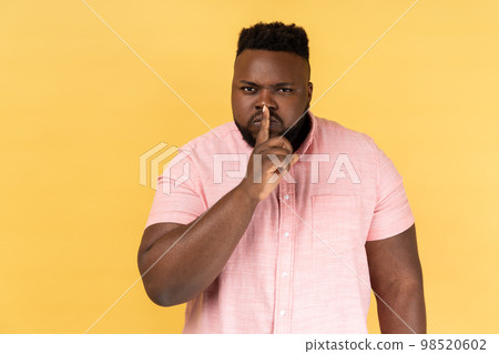 Shh, make silence please. Portrait of serious strict man wearing pink shirt standing showing quiet gesture, saying hush, keep secret. Indoor studio shot isolated on yellow background. 98520602
