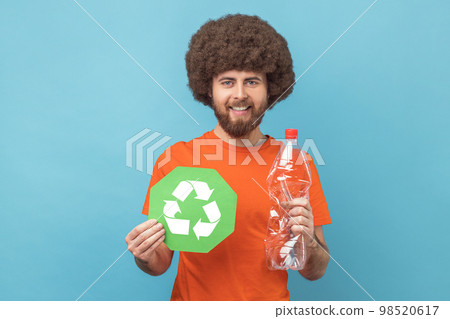 Portrait of smiling man with Afro hairstyle wearing orange T-shirt holding recycling green sign and plastic bottle, looking at camera. Indoor studio shot isolated on blue background. 98520617