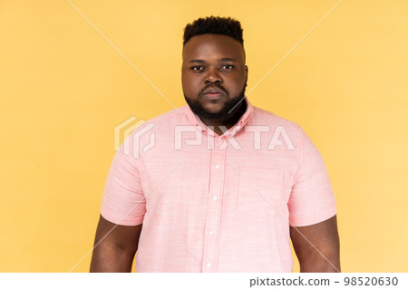Portrait of serious strict young adult man wearing pink shirt standing and looking at camera with bossy facial expression, being in bad mood. Indoor studio shot isolated on yellow background. Portrait of serious strict young adult man wearing pink shirt standing and looking at camera with bossy facial expression, being in bad mood. Indoor studio shot isolated on yellow background. 98520630