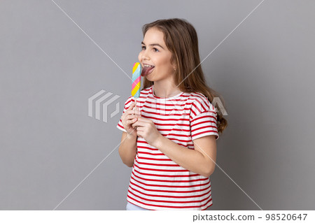 Portrait of little girl wearing striped T-shirt holding colorful candy, licking, looking away with childish satisfied expression. Indoor studio shot isolated on gray background. Portrait of little girl wearing striped T-shirt holding colorful candy, licking, looking away with childish satisfied expression. Indoor studio shot isolated on gray background. 98520647