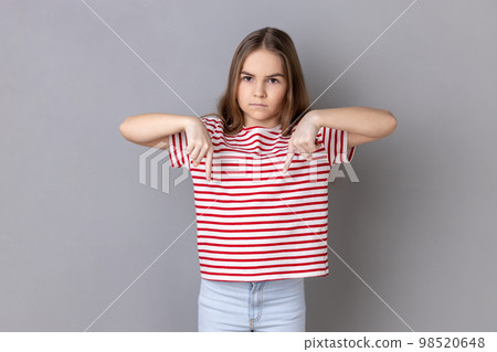 Here and right now. Portrait of serious bossy dark haired little girl wearing striped T-shirt standing and pointing index fingers down. Indoor studio shot isolated on gray background. Here and right now. Portrait of serious bossy dark haired little girl wearing striped T-shirt standing and pointing index fingers down. Indoor studio shot isolated on gray background. 98520648