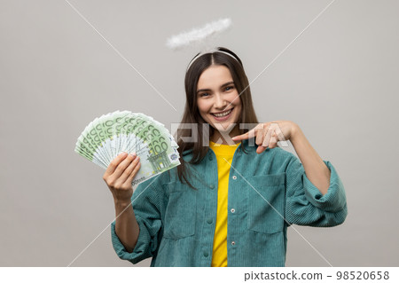 Cheerful dark haired woman with halo on head pointing finger on bunch of euro banknotes and smiling, profit, wearing casual style jacket. Indoor studio shot isolated on gray background. 98520658