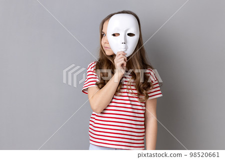 Portrait of dark haired little cute girl wearing striped T-shirt holding white mask and looking away with serious facial expression. Indoor studio shot isolated on gray background. Portrait of dark haired little cute girl wearing striped T-shirt holding white mask and looking away with serious facial expression. Indoor studio shot isolated on gray background. 98520661