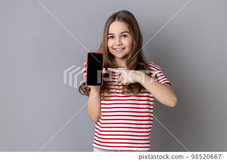 Portrait of delighted little girl wearing striped T-shirt pointing finger at smartphone with white empty display, bragging with application. Indoor studio shot isolated on gray background. Portrait of delighted little girl wearing striped T-shirt pointing finger at smartphone with white empty display, bragging with application. Indoor studio shot isolated on gray background. 98520667