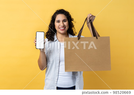 Portrait of smiling happy woman with dark wavy hair holding paper shopping bag and smart phone with empty display for advertisement. Indoor studio shot isolated on yellow background. 98520669