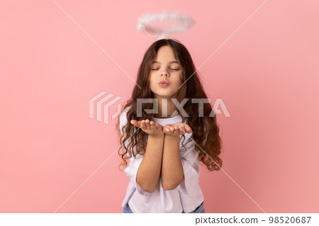 Portrait of little girl wearing white T-shirt with halo on head sending air kiss on camera, flirting, valentines day, keeps eyes closed. Indoor studio shot isolated on pink background. 98520687