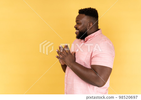 Side view portrait of cunning bearded man wearing pink shirt clasping hands and planning evil tricky prank or scheming, cheating with sly smile. Indoor studio shot isolated on yellow background. Side view portrait of cunning bearded man wearing pink shirt clasping hands and planning evil tricky prank or scheming, cheating with sly smile. Indoor studio shot isolated on yellow background. 98520697
