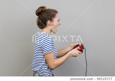Side view portrait of woman wearing striped T-shirt holding gamepad, winning video games competition, screaming happy expression. Indoor studio shot isolated on gray background. 98520727