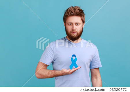 Portrait of serious young adult attractive bearded man holding blue awareness, disease symbol, looking at camera, support and care. Indoor studio shot isolated on blue background. 98520772