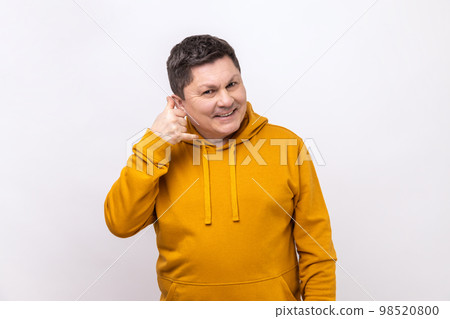 Portrait of handsome cheerful man holding telephone hand gesture near ear, imitating communication on phone, wearing urban style hoodie. Indoor studio shot isolated on white background. 98520800