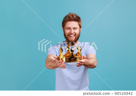 Portrait of smiling happy bearded man holding out golden crown, looking at camera with toothy smile, expressing positive emotions. Indoor studio shot isolated on blue background. 98520842