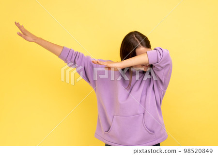 Winning, success gesture. Portrait of woman showing dab dance pose, famous internet meme, performing dabbing trends, wearing purple hoodie. Indoor studio shot isolated on yellow background. 98520849