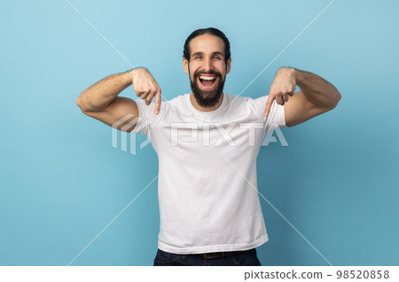 Look at ads below. Portrait of man with beard wearing white T-shirt smiling and pointing down, showing place for idea presentation, commercial text. Indoor studio shot isolated on blue background. 98520858