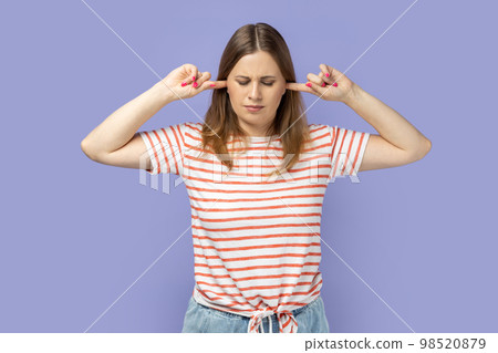 Portrait of depressed attractive blond woman wearing striped T-shirt standing covering ears with fingers, being irritated by noise, closed her eyes. Indoor studio shot isolated on purple background. 98520879
