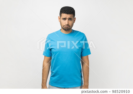 Portrait of sad upset depressed unshaven man wearing blue T- shirt standing looking at camera with frowning face, expressing sadness, being offended. Indoor studio shot isolated on gray background. Portrait of sad upset depressed unshaven man wearing blue T- shirt standing looking at camera with frowning face, expressing sadness, being offended. Indoor studio shot isolated on gray background. 98520910
