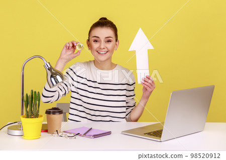 Portrait of happy optimistic woman holding golden bitcoin and white paper arrow, showing growth of crypto currency, sitting on workplace. Indoor studio studio shot isolated on yellow background. 98520912