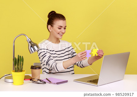 Satisfied woman showing to laptop display two colored pieces of jigsaw, easily solving difficult tasks at work, sitting at workplace. Indoor studio studio shot isolated on yellow background. 98520917