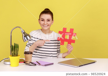 Young attractive woman sitting at workplace, pointing at big red hashtag symbol to laptop screen, sharing viral content, tagged message. Indoor studio studio shot isolated on yellow background. 98520924