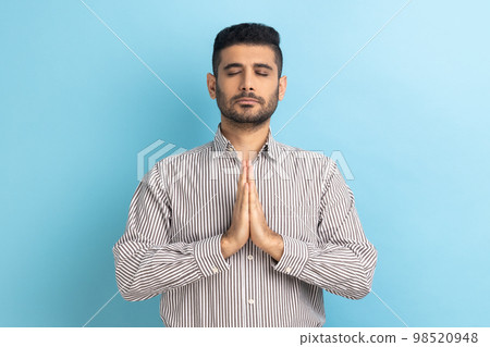 Portrait of calm relaxed young businessman with beard standing doing yoga meditating exercise, keeping palms together, wearing striped shirt. Indoor studio shot isolated on blue background. 98520948