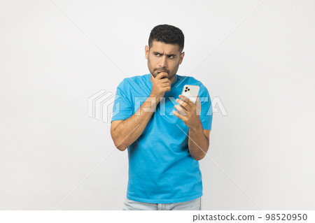 Portrait of pensive thoughtful unshaven man blogger wearing blue T- shirt standing using cell phone, writing post, thinking about answer the message. Indoor studio shot isolated on gray background. 98520950