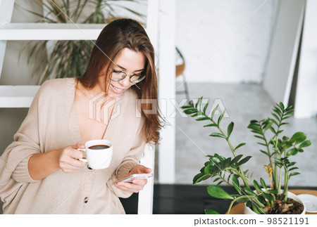 Smiling woman with long hair in beige cardigan drinking coffe and using mobile phone at home Smiling woman with long hair in beige cardigan drinking coffe and using mobile phone at home 98521191