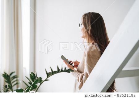 Smiling woman with long hair in beige cardigan drinking coffe and using mobile phone at home Smiling woman with long hair in beige cardigan drinking coffe and using mobile phone at home 98521192
