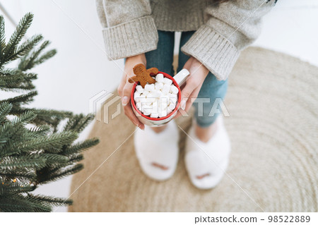Young woman in cozy sweater with cup of cocoa with marshmallow in room with Christmas tree at home 98522889