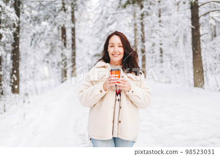 Smiling woman with paper cup of coffee in hands against background of snowy winter forest Smiling woman with paper cup of coffee in hands against background of snowy winter forest 98523031