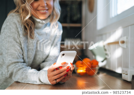 Young woman with blonde curly hair in grey sweater using mobile phone in hands in kitchen at house 98523734