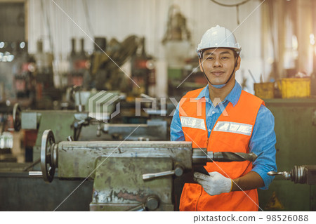 portrait Asian engineer adult male worker in machine shop with check list happy smile in safety hardhat 98526088
