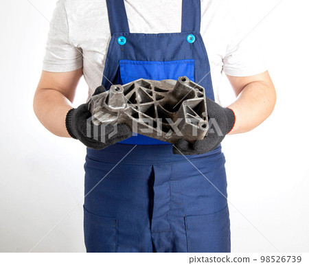 A car mechanic in blue overalls holds a pillow of a car engine in his hands. The concept of replacing an aluminum engine mount in a car, an automotive spare part. Close-up A car mechanic in blue overalls holds a pillow of a car engine in his hands. The concept of replacing an aluminum engine mount in a car, an automotive spare part. Close-up 98526739