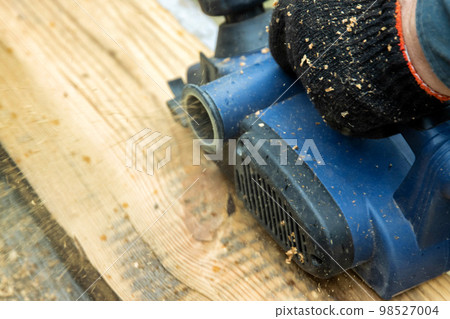 A male carpenter plans boards with an electric planer to make a bench. Carpentry, industry. Woodworker A male carpenter plans boards with an electric planer to make a bench. Carpentry, industry. Woodworker 98527004