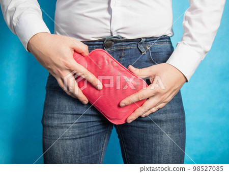 A man holds a red heating pad with water near his groin on a blue background. The concept of treatment of trauma to the scrotum and prostatitis, close-up A man holds a red heating pad with water near his groin on a blue background. The concept of treatment of trauma to the scrotum and prostatitis, close-up 98527085